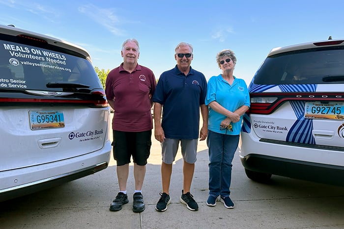 Three volunteer drivers with Valley Senior Services smile and stand between two delivery vehicles donated by Gate City Bank