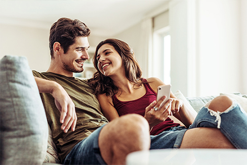 Young couple on sofa, smiling while comparing different CD rates and specials from Gate City Bank on a smartphone