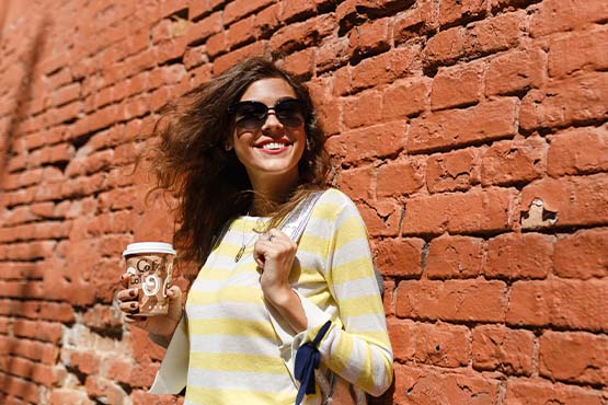 Young brunette in sunglasses, holding a cup of coffee while leaning up against a historic building in Elk River, MN