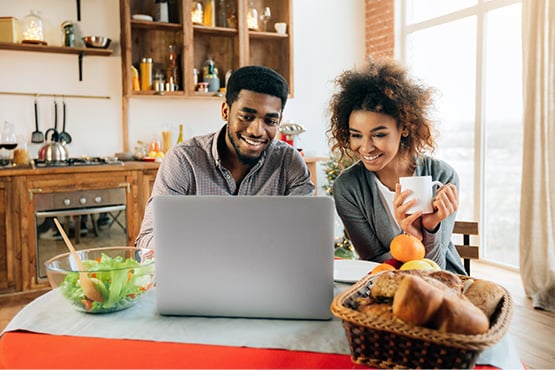 a happy couple sits up to a laptop on their kitchen table to learn Gate City Bank’s tips for saving on home closing costs