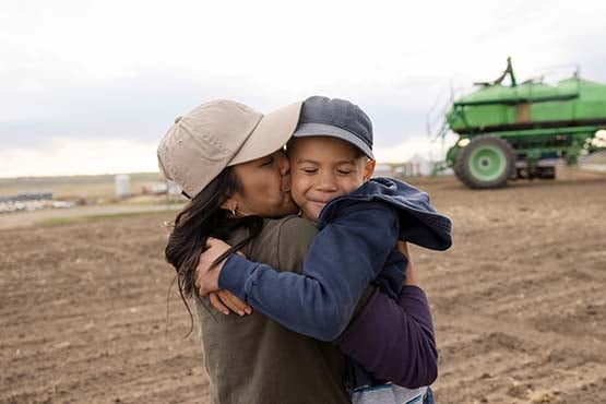 Smiling boy in a baseball cap, getting a bear hug from his mom on their farm outside Wahpeton, ND